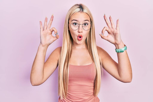 Young blonde girl wearing casual clothes looking surprised and shocked doing ok approval symbol with fingers. crazy expression  - Foto, immagini