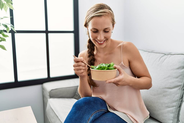 Молода біла жінка посміхається впевнено їсть салат вдома
 - Фото, зображення