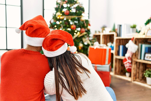 Young latin couple on back view sitting on the floor looking christmas tree at home. - Photo, image