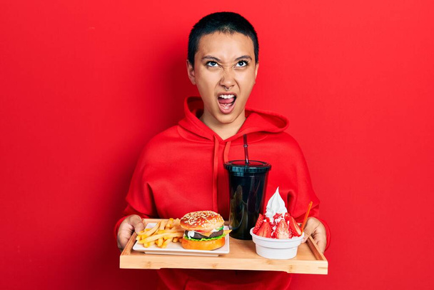 Krásná hispánská žena s krátkými vlasy jíst chutný klasický burger s hranolky a limonádou naštvaný a šílený křičí frustrovaný a zuřivý, křičí vzteky vzhlíží.  - Fotografie, Obrázek
