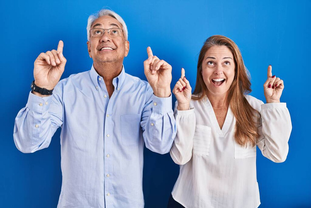 Middelbare leeftijd hispanic paar staan over blauwe achtergrond glimlachend verbaasd en verrast en wijzen met vingers en opgeheven armen.  - Foto, afbeelding