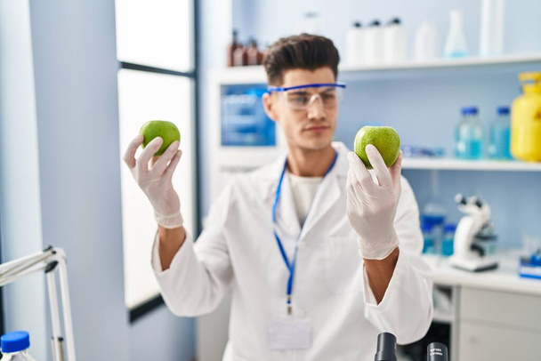 Young hispanic man wearing scientist uniform holding apples at laboratory - Photo, image