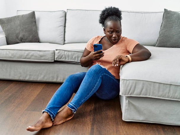 Mladá africká žena používající smartphone sedí doma na podlaze a dívá se na hodinky a bojí se, že se opozdí  - Fotografie, Obrázek