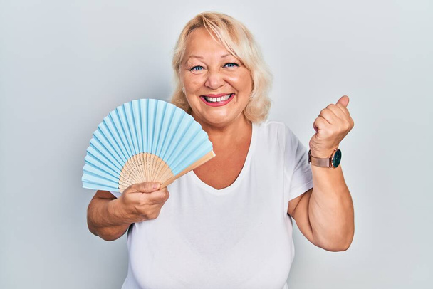 Middle age blonde woman waving hand fan cooling air in summer screaming proud, celebrating victory and success very excited with raised arm  - Φωτογραφία, εικόνα