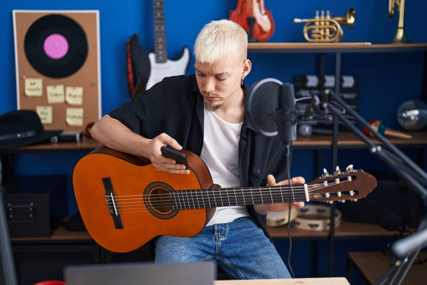 Young caucasian man musician playing classical guitar using smartphone at music studio - 写真・画像