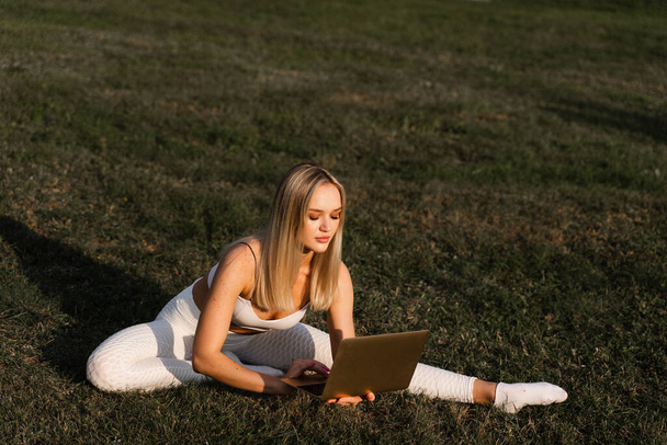 Entraînement de fitness en ligne avec ordinateur portable dans le parc. Fit blonde assise sur l'herbe et bavarder avec le formateur. Style de vie sportif extérieur - Photo, image