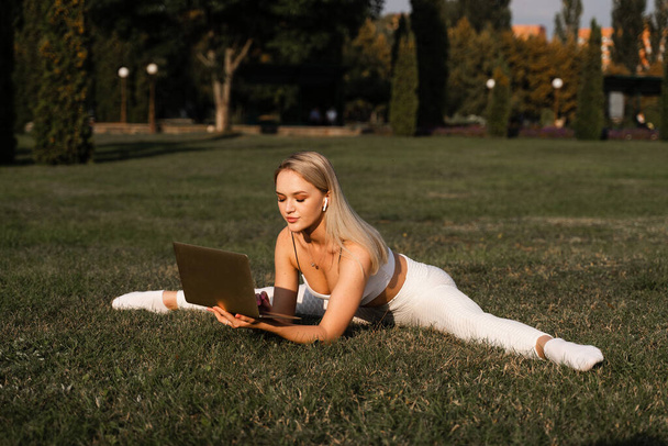 Fit blonde doet been gespleten op het gras. Online stretching training met laptop in het park. Sport levensstijl - Foto, afbeelding