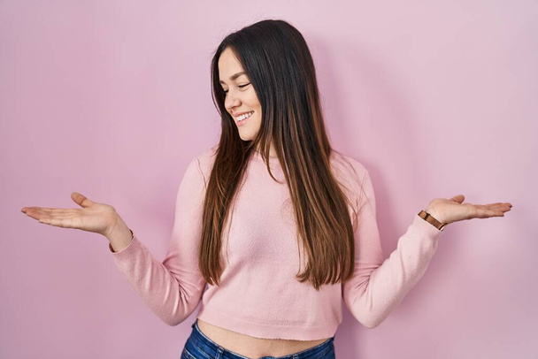 Jeune femme brune debout sur fond rose souriant montrant les paumes ouvertes des deux mains, présentant et annonçant comparaison et équilibre  - Photo, image