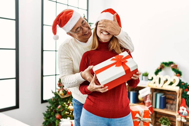 Mediana edad pareja hispana sonriendo feliz y abrazando celebración de regalo de Navidad en casa. - Foto, imagen