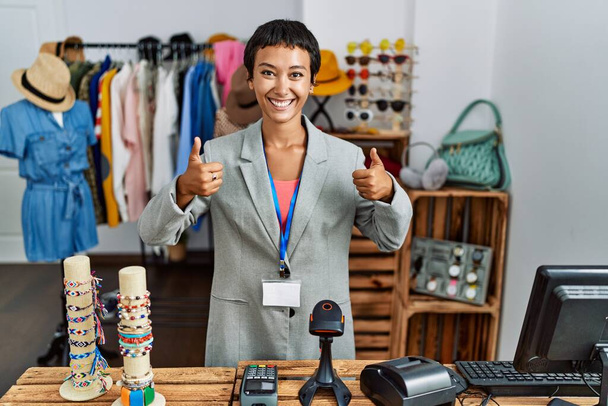 Молода іспаномовна жінка з коротким волоссям працює менеджером в роздрібному магазині успіху знак робить позитивний жест рукою, великі пальці вгору посміхаються і щасливі. веселий вираз і жест переможця
.  - Фото, зображення
