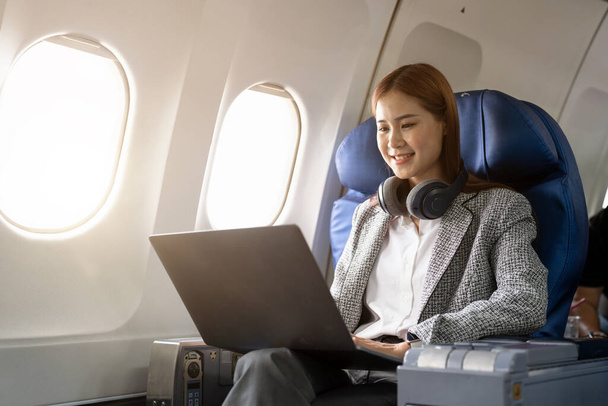 飛行機のファーストクラスで窓の近くに座ってラップトップを使用してヘッドフォンを身に着けているアジアの若い女性,旅行やビジネスコンセプト. - 写真・画像