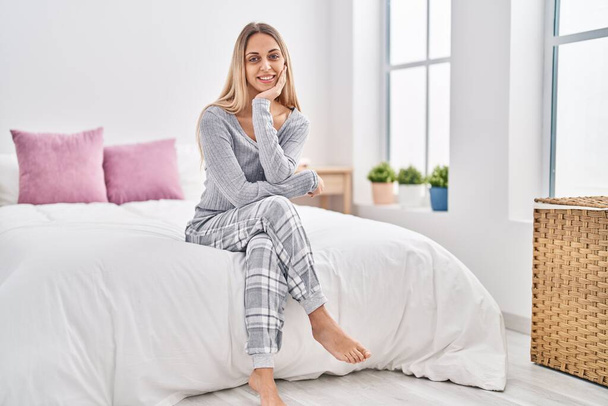 Mujer joven sonriendo confiada sentada en la cama en el dormitorio - Foto, Imagen