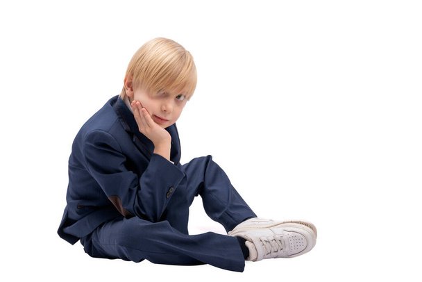 Blond boy in school uniform sits on white background. Portrait of student of the 1st or 2nd grade. - Photo, Image