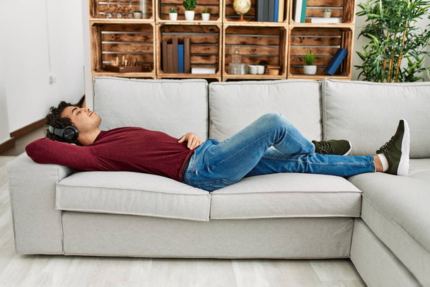 Junger hispanischer Mann hört entspannt Musik, die zu Hause auf dem Sofa liegt. - Foto, Bild
