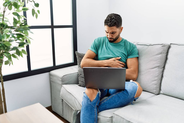 Νέος όμορφος άντρας με γενειάδα χρησιμοποιεί φορητό υπολογιστή κάθεται στον καναπέ στο σπίτι με το χέρι στο στομάχι, επειδή ναυτία, επώδυνη ασθένεια αίσθημα αδιαθεσίας. έννοια του πόνου.  - Φωτογραφία, εικόνα