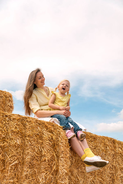Retrato de una joven madre feliz con una hija pequeña sentada en un pajar en la granja. La familia se divierte. Cosecha de heno - Foto, Imagen