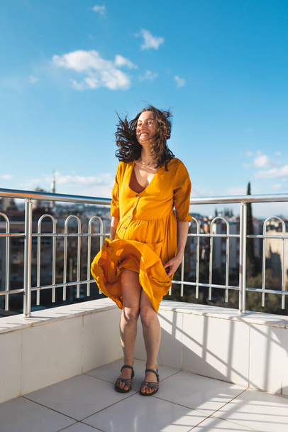 Egy sárga ruhás lány áll az erkélyen egy erős szélben, egy mosolygó nő áll a tetőn, kilátással a városra, és a szél felfújja a ruháját, egy vonzó lány néz a városra - Fotó, kép