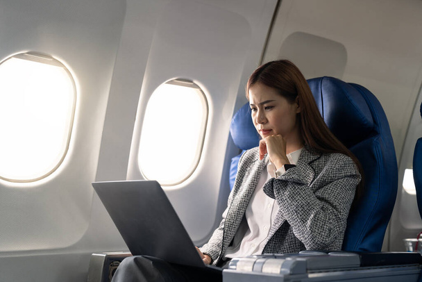 Ασιάτισσα νεαρή γυναίκα που χρησιμοποιεί φορητό υπολογιστή κάθεται κοντά σε παράθυρα στην πρώτη θέση στο αεροπλάνο κατά τη διάρκεια της πτήσης, Ταξίδια και Επιχειρήσεων έννοια. - Φωτογραφία, εικόνα