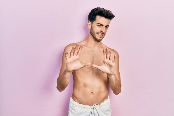 Junge hispanische Mann in Badebekleidung ohne Hemd angewidert Ausdruck, unzufrieden und ängstlich tun Ekel Gesicht, weil Abneigung Reaktion. mit erhobenen Händen  - Foto, Bild