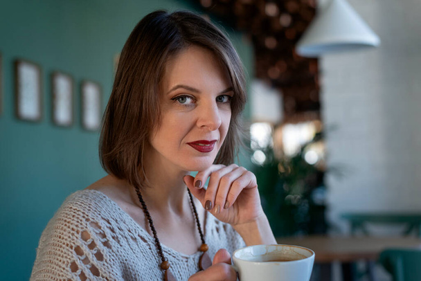 Portrait of middle-aged woman drinks coffee on inside cafe. Portrait of brunette girl sits at table in cafe - Photo, image