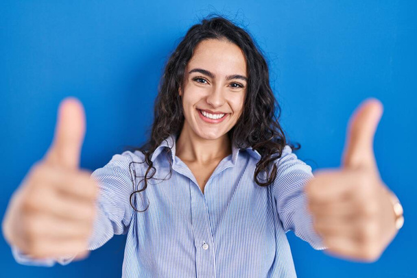 Young brunette woman standing over blue background approving doing positive gesture with hand, thumbs up smiling and happy for success. winner gesture.  - Foto, immagini