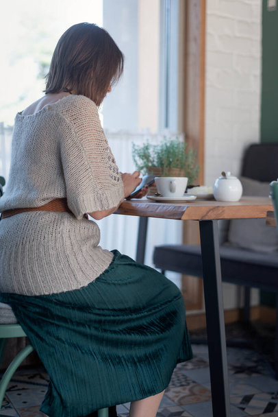 Woman in long skirt sits at table in cafe with cup of coffee or tea and telephone. Vertical frame - Photo, Image