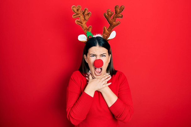 Young hispanic woman wearing deer christmas hat and red nose shouting suffocate because painful strangle. health problem. asphyxiate and suicide concept.  - Photo, image