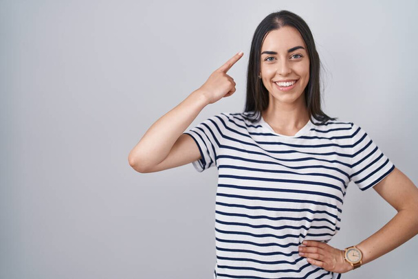 Mladá brunetka žena v pruhované tričko s úsměvem ukazuje na hlavu s jedním prstem, skvělý nápad nebo myšlenka, dobrá paměť  - Fotografie, Obrázek