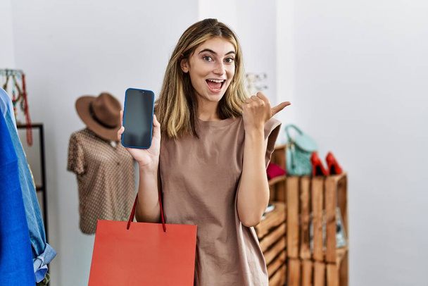Młoda blondynka trzyma torby na zakupy pokazując ekran smartfona wskazując kciuk w górę do boku uśmiechnięta szczęśliwa z otwartymi ustami  - Zdjęcie, obraz