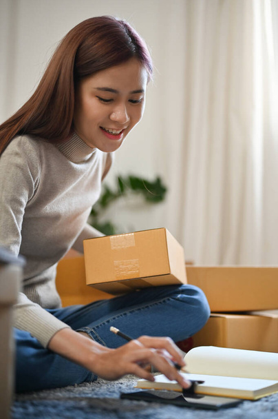 Portrait, Beautiful female online shop owner checking her customer's address on the phone and preparing a parcel box. SME business - 写真・画像