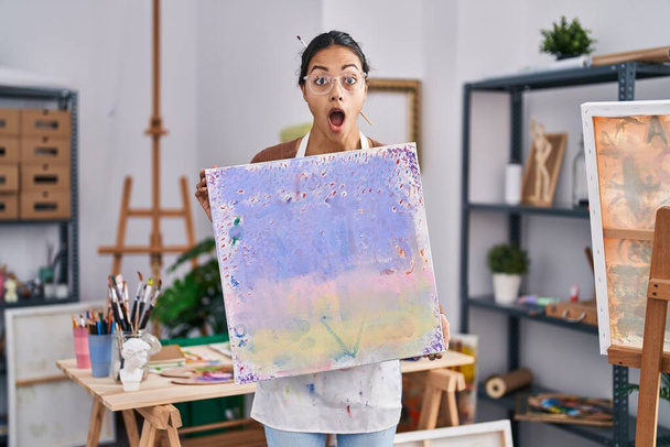 Jonge Braziliaanse vrouw houdt schildersdoek in kunststudio bang en geschokt met verbazing en verbaasde uitdrukking, angst en opgewonden gezicht.  - Foto, afbeelding