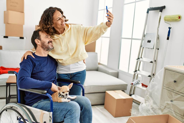 Das hispanische Paar mittleren Alters lächelt glücklich. Mann sitzt im Rollstuhl mit Hund an den Beinen und Frau macht Selfie mit dem Smartphone im neuen Zuhause. - Foto, Bild