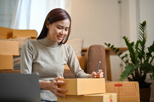 Úspěšné mladé asijské žena on-line obchod majitel připravuje přepravní objednávky, psaní adresy zákazníka na krabici v jejím obývacím pokoji. - Fotografie, Obrázek