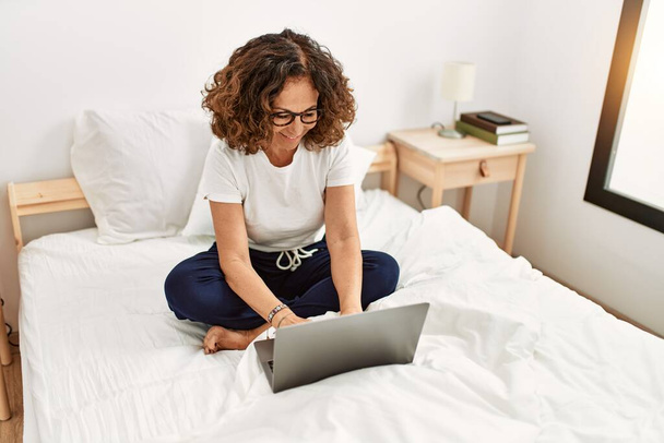 Orta yaşlı İspanyol kadın yatak odasında dizüstü bilgisayar kullanarak gülümsüyor. - Fotoğraf, Görsel