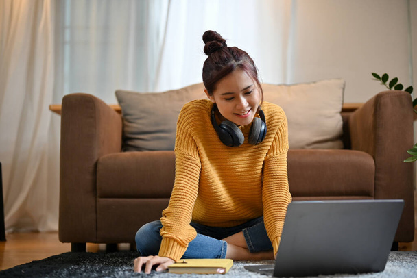 Entspannte und glückliche Asiatin sitzt mit tragbarem Laptop auf einem Teppich im Wohnzimmer und schaut sich etwas im Internet an. - Foto, Bild