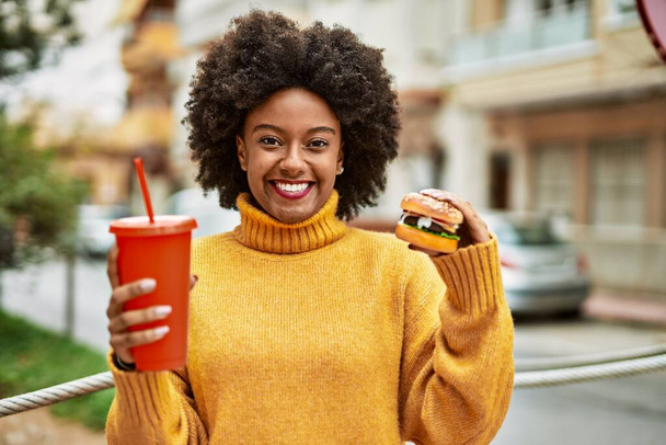 Молодая африканская американка ест классический бургер и пьет газировку в городе. - Фото, изображение
