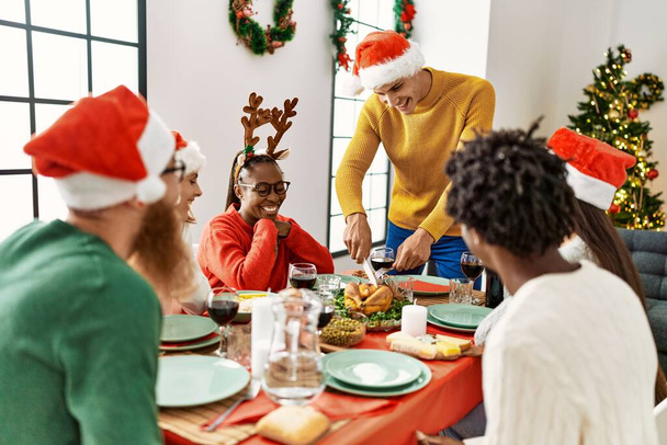 Grupa młodych ludzi uśmiechnięty szczęśliwy mając świąteczny obiad, Człowiek cięcia pieczonego indyka służyć w domu. - Zdjęcie, obraz