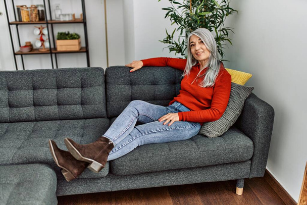 Donna dai capelli grigi di mezza età sorridente fiduciosa seduta sul divano di casa - Foto, immagini