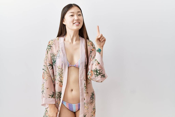 Mladá asijská žena nosí bikiny a květinové kimono ukazující a ukazující nahoru s prstem číslo jedna při úsměvu sebevědomé a šťastné.  - Fotografie, Obrázek