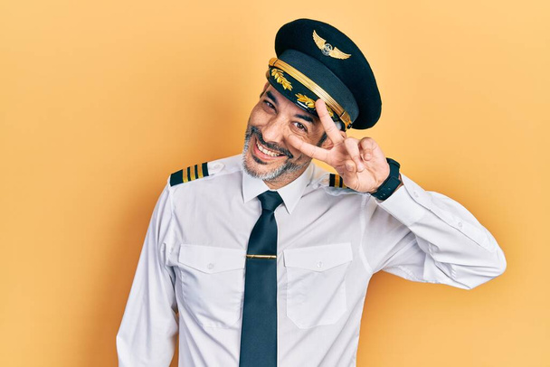 Handsome middle age man with grey hair wearing airplane pilot uniform doing peace symbol with fingers over face, smiling cheerful showing victory  - Photo, image