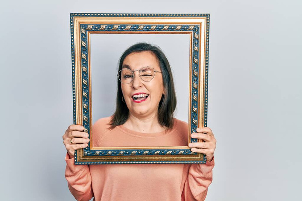Middle age hispanic woman holding empty frame winking looking at the camera with sexy expression, cheerful and happy face.  - 写真・画像