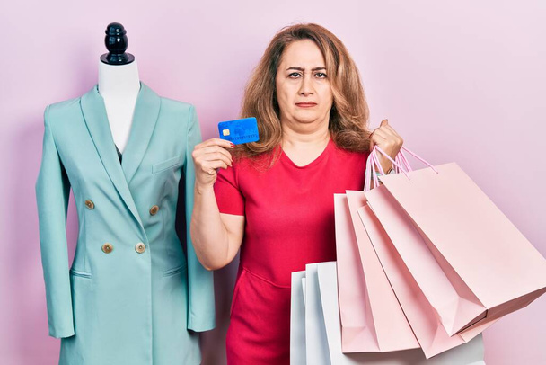 Orta yaşlı beyaz bir kadın alışveriş poşetleri tutuyor ve kredi kartı konusunda şüpheci ve gergin, sorun yüzünden suratını asıyor. negatif kişi.  - Fotoğraf, Görsel