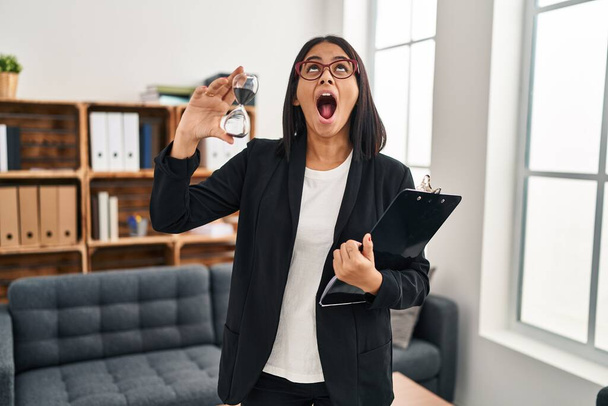 Young hispanic woman working at consultation office holding sand clock angry and mad screaming frustrated and furious, shouting with anger looking up.  - Photo, Image