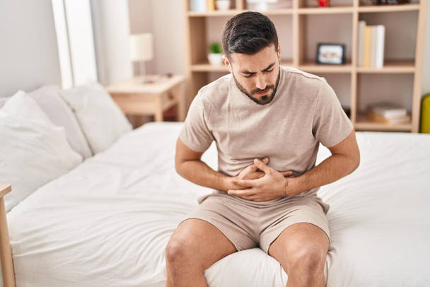 Young hispanic man suffering for stomachache sitting on bed at bedroom - Photo, image