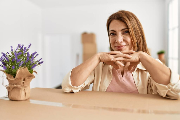 Middle age caucasian woman smiling happy leaning on cardboard box at new home. - 写真・画像