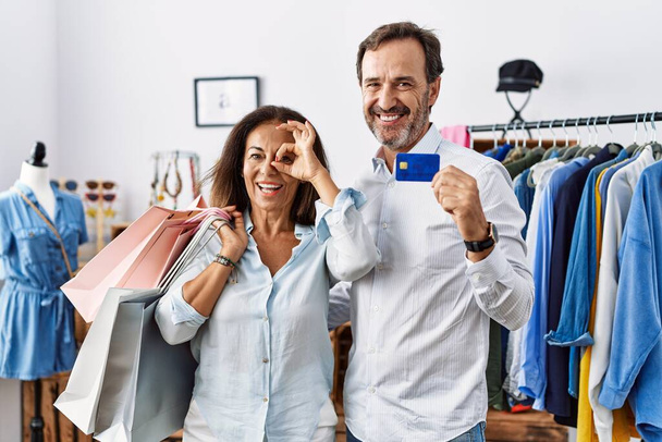 Латиноамериканская пара среднего возраста, держащая сумки с покупками и кредитную карту, делает все хорошо жест с улыбкой на руке, глаза смотрят сквозь пальцы со счастливым лицом.  - Фото, изображение