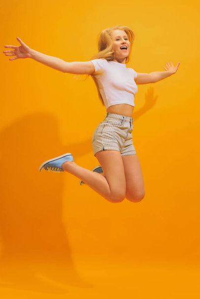 Jump for joy. Emotional young girl with long blond hair wearing white t-shirt posing isolated on yellow background. Concept of human emotions, facial expression, youth, aspiration, ad - Fotoğraf, Görsel