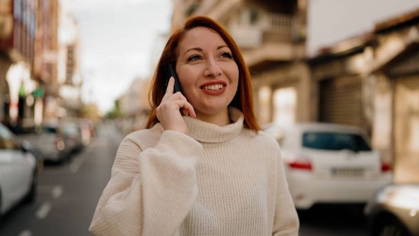 Νεαρή κοκκινομάλλα γυναίκα χαμογελά αυτοπεποίθηση μιλώντας στο smartphone στο δρόμο - Φωτογραφία, εικόνα