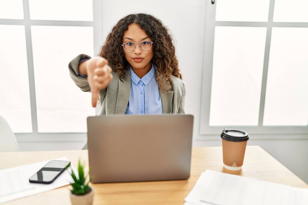 Beautiful hispanic business woman sitting on desk at office working with laptop looking unhappy and angry showing rejection and negative with thumbs down gesture. bad expression.  - Photo, Image
