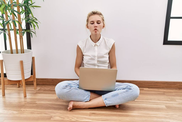 Young blonde woman using computer laptop sitting on the floor at the living room making fish face with lips, crazy and comical gesture. funny expression.  - Foto, Imagem